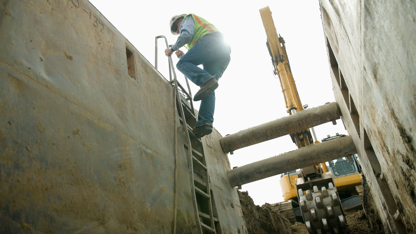 Worker in safety vest climbing out of a deep shored excavation beside heavy equipment